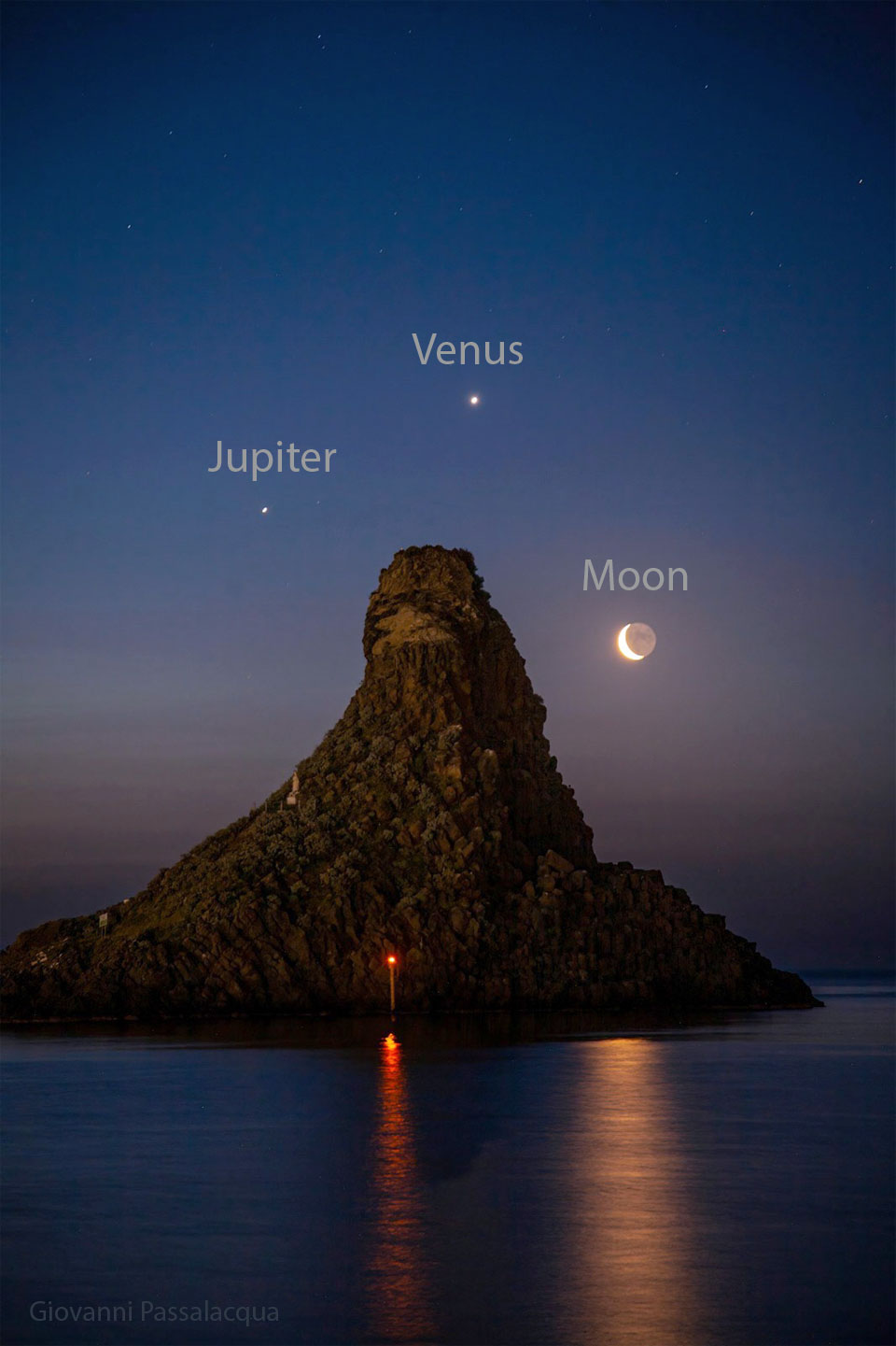 A seascape surrounds a large tree-covered hill. Surrounding the hill
in the night sky are three bright dots: the planets Jupiter, Venus,
and a crescent Moon.
Please see the explanation for more detailed information.