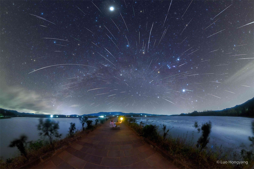 Na sliki sta dve osebi, ki zreta v temno,
z zvezdami napolnjeno nebo. Nebo je prekrito s sledmi, ki 
jih povzroÄajo meteorji roja Geminidov.
Za podrobnejĹĄe informacije gle razlago.