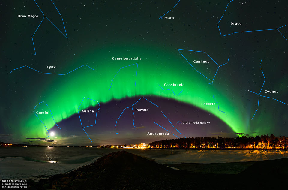 
A big green arc is seen arching across the night sky. The arc
fades away above into a green haze, while no green glow is seen
below the arc. A dark sky filled with stars and constellations
fills the background. Snow and distant trees line the foreground.
Please see the explanation for more detailed information.