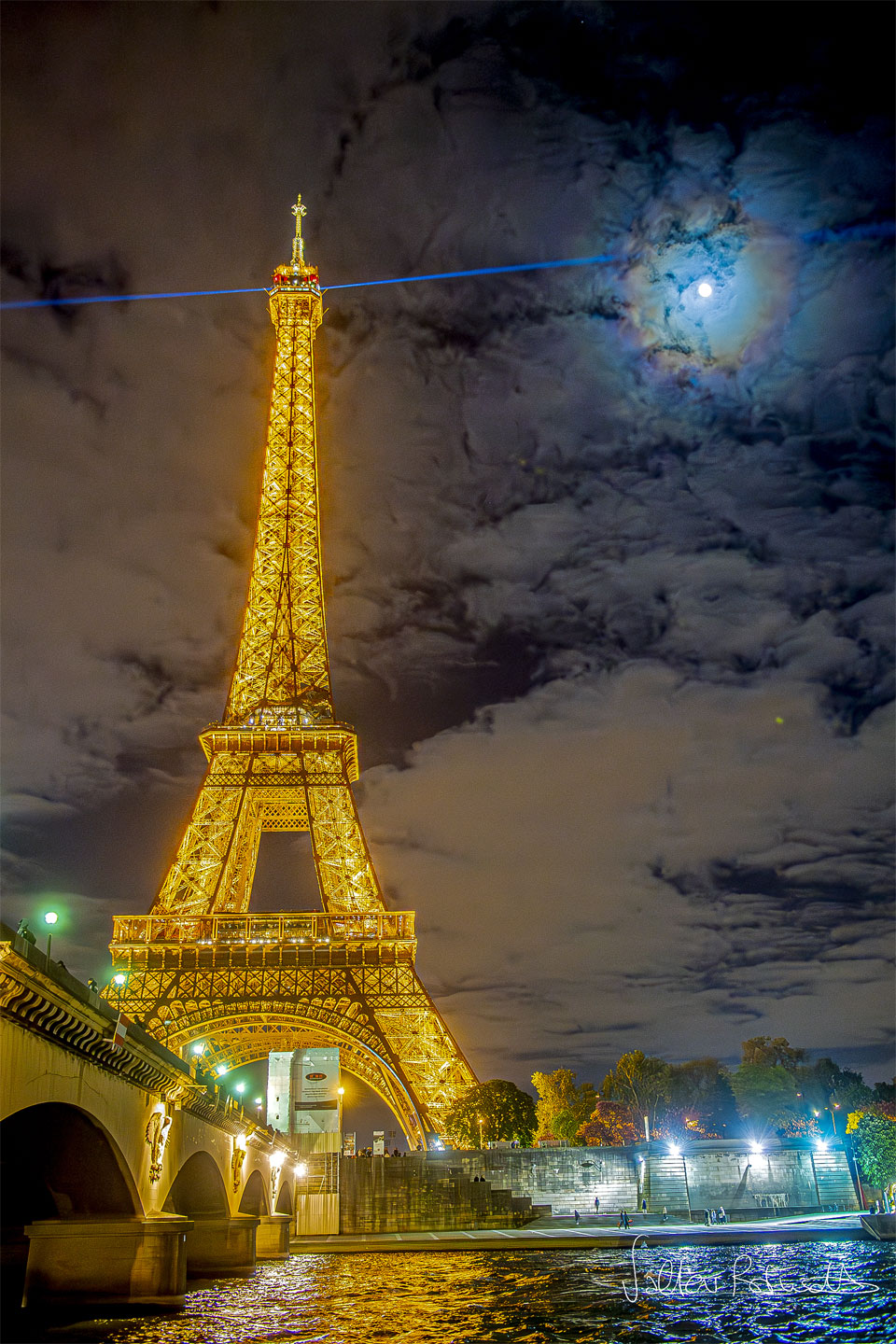 The famous Eiffel Tower in Paris, France is pictured
on the left lit up in gold at night. A blue laser shines
out from the top. Clouds dot the background sky. The Moon
is also visible through the clouds, but is circled by
colorful rings: a lunar corona.
Please see the explanation for more detailed information.