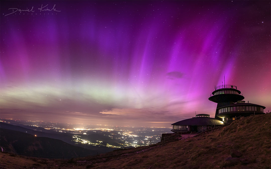 Purple striped aurora cover a star filled sky. Mountain
peaks are visible on the sides, as well as a futuristic
looking building on the right side. City lights are
visible in the valley down below.
Please see the explanation for more detailed information.