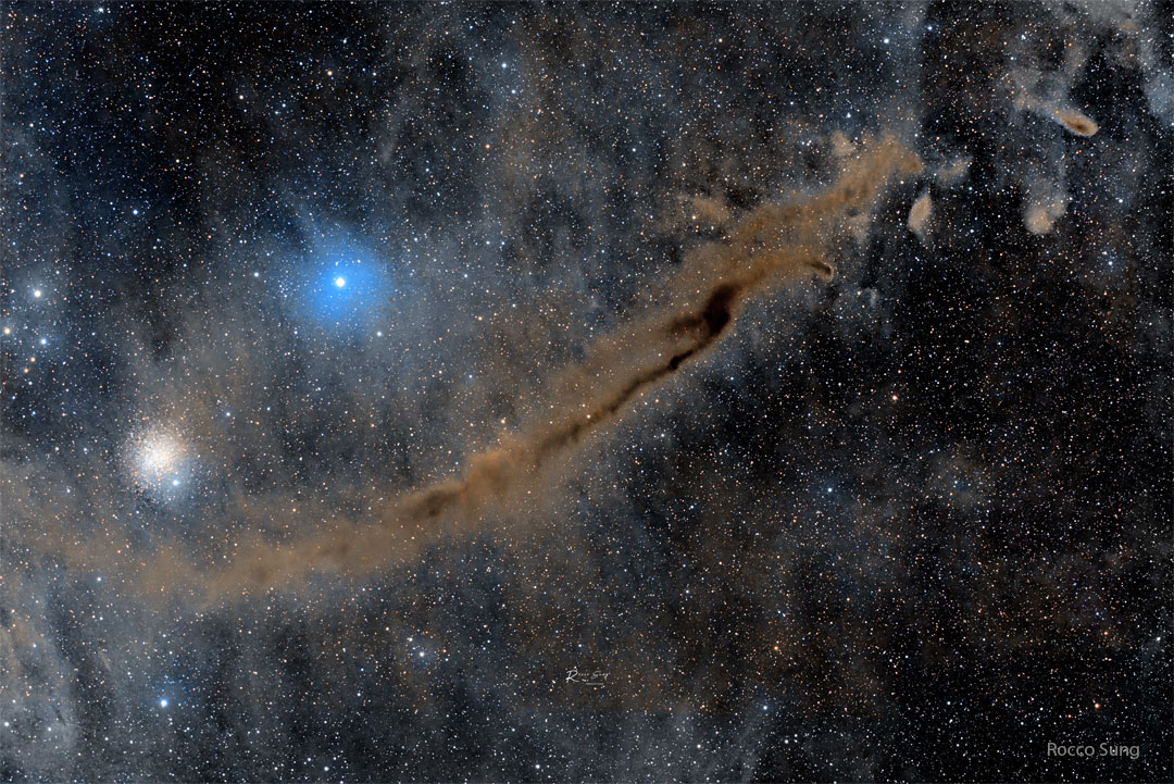 A busy starfield is shown which an elongated brown
nebula running diagonally from the lower left to the
upper right. A bright blue star and a star cluster
appear above the nebula.
Please see the explanation for more detailed information.