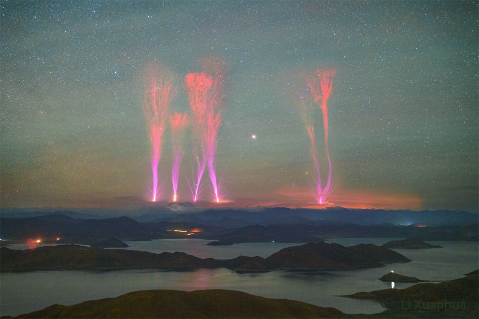 A landscape showing a night sky over distant mountains
is shown. Lakes dot the foreground in front of the mountains.
Extending from above the mountains into the night sky are
six bright jets. The jets are violet at the bottom but red
at the top.
Please see the explanation for more detailed information.