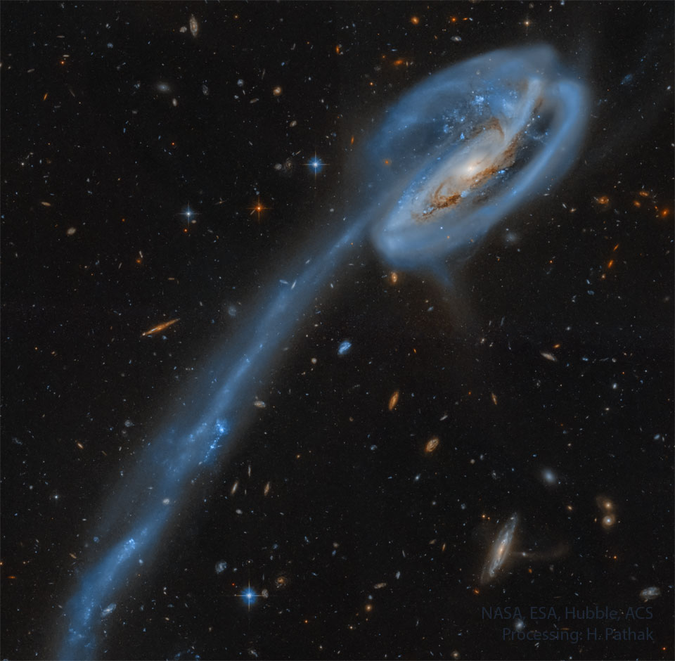 A spiral galaxy is shown on the upper left with a really long tail of stars and blue-glowing gas trailing to the lower left. Please see the explanation for more detailed information.