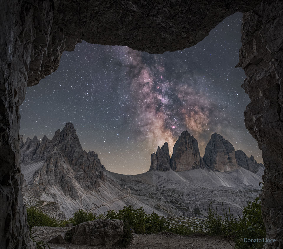 A starry sky is seen through a rectangular rock
opening. Three rocky peaks appear toward the right, while
other peaks appear toward the left. Rising above the
right peaks is the central band of our Milky Way Galaxy.
Please see the explanation for more detailed information.