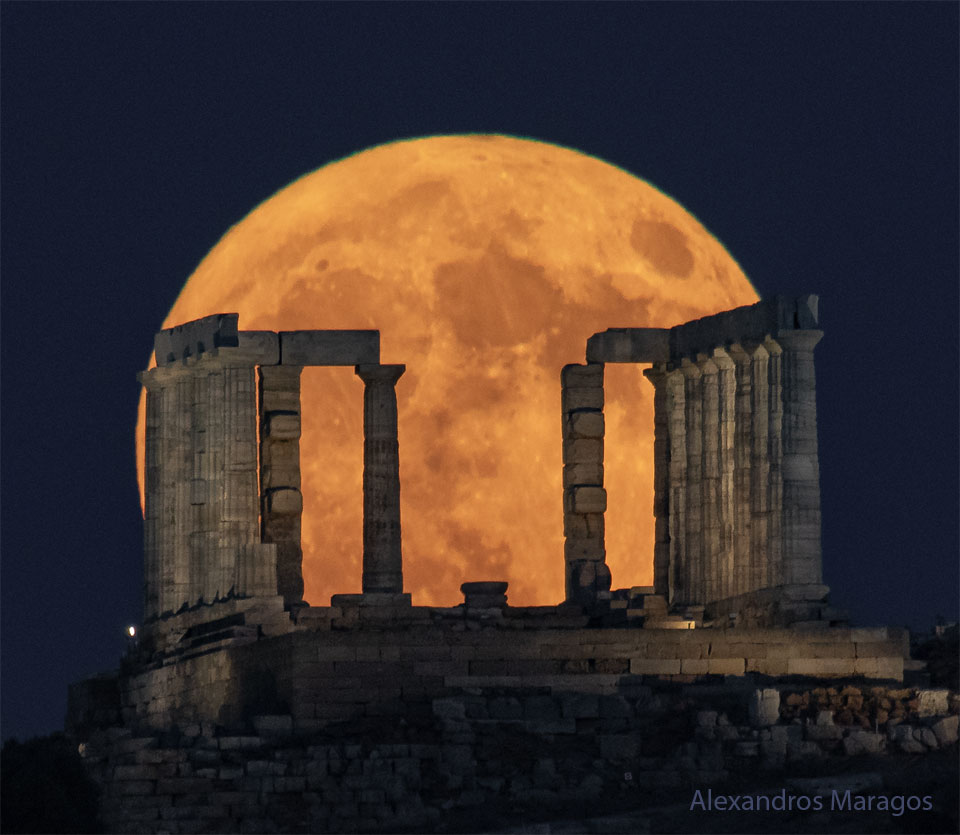 A large and orange-tinted moon is pictured rising beyond
the pillars of an ancient structure. The foreground is dark and
the night sky behind the Moon appear blue.
Please see the explanation for more detailed information.