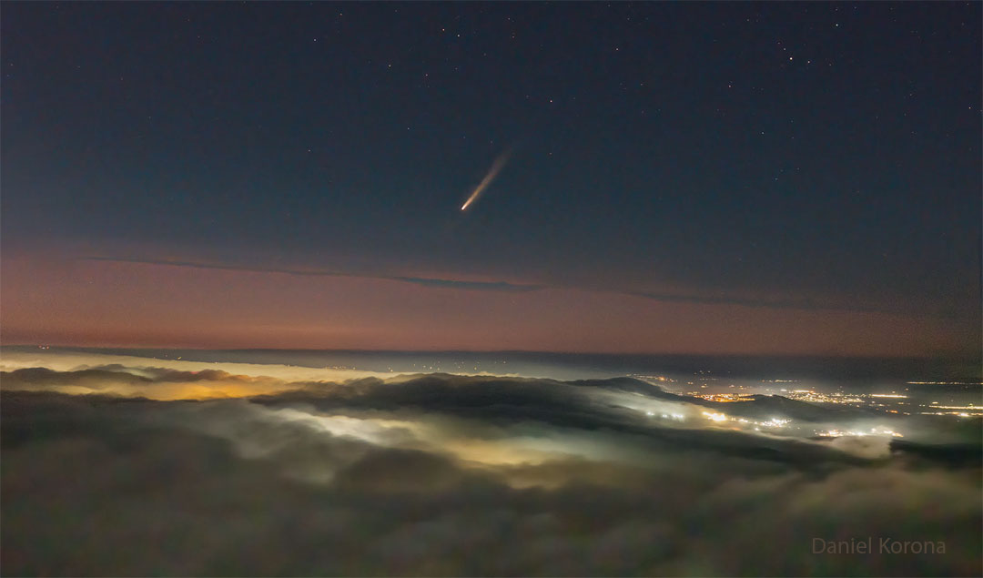 A picture shows clouds across the bottom and a dark night sky
across the top. In the middle is a band of orange sky. City lights
are visible on the right through gaps in the clouds. In the center
of the upper sky is a comet with its tail pointing toward the
upper right.
Please see the explanation for more detailed information.