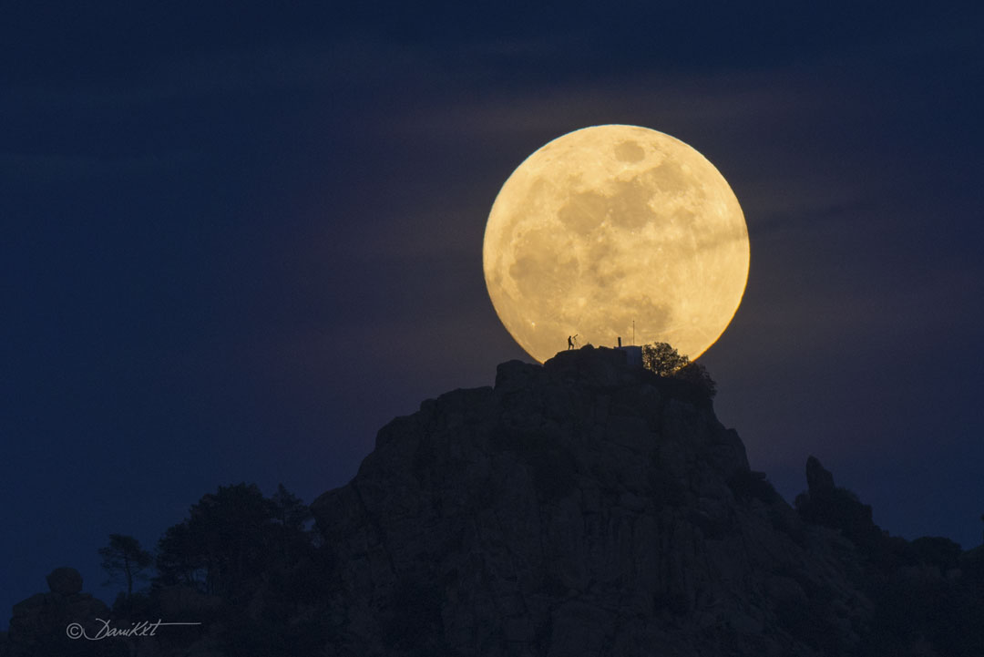 Earth's Moon is shown just beyond a rocky hill. The Moon is
near full phase. On the hill the silhouette of a person looking
through a telescope can be seen. A rollover darkens part of the
Moon that looks to some like a human face.
Please see the explanation for more detailed information.