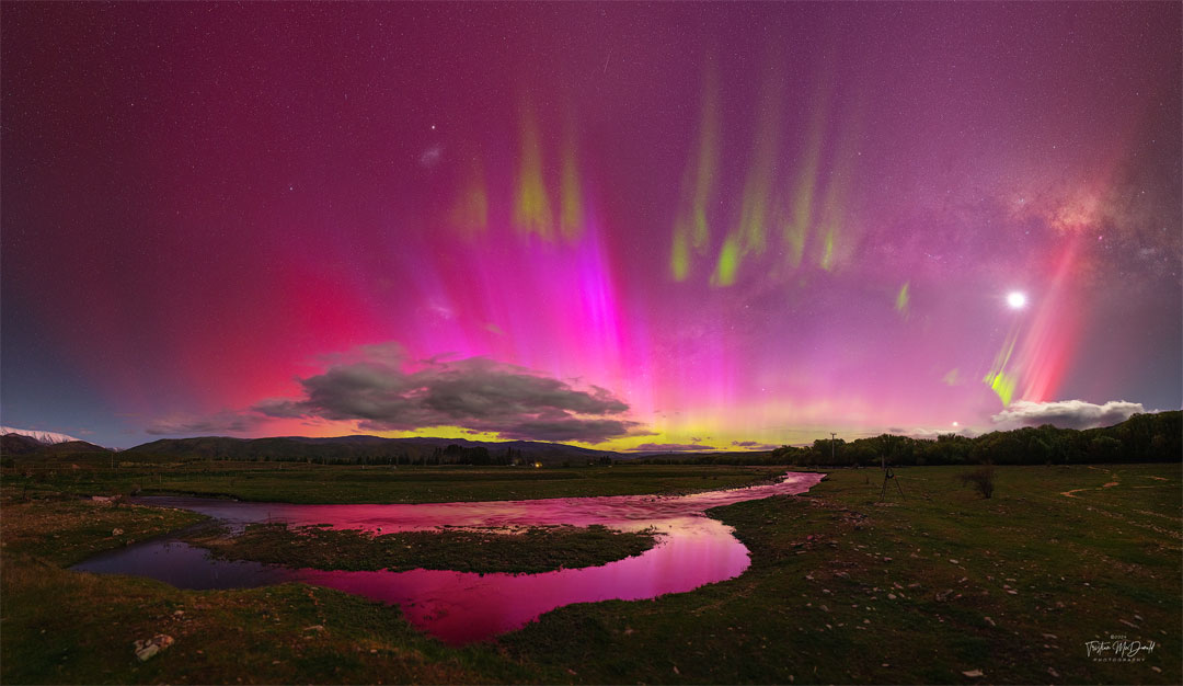 A night sky is shown that appears mostly red due to pervasive
aurora. In the foreground is covered by watery grasslands. Clouds
are visible above the horizon. Thin green aurora are visible
toward the top of the frame. In the background one can find the Moon,
the LMC, SMC, Venus, a meteor, and the band of our Milky Way galaxy.
Please see the explanation for more detailed information.