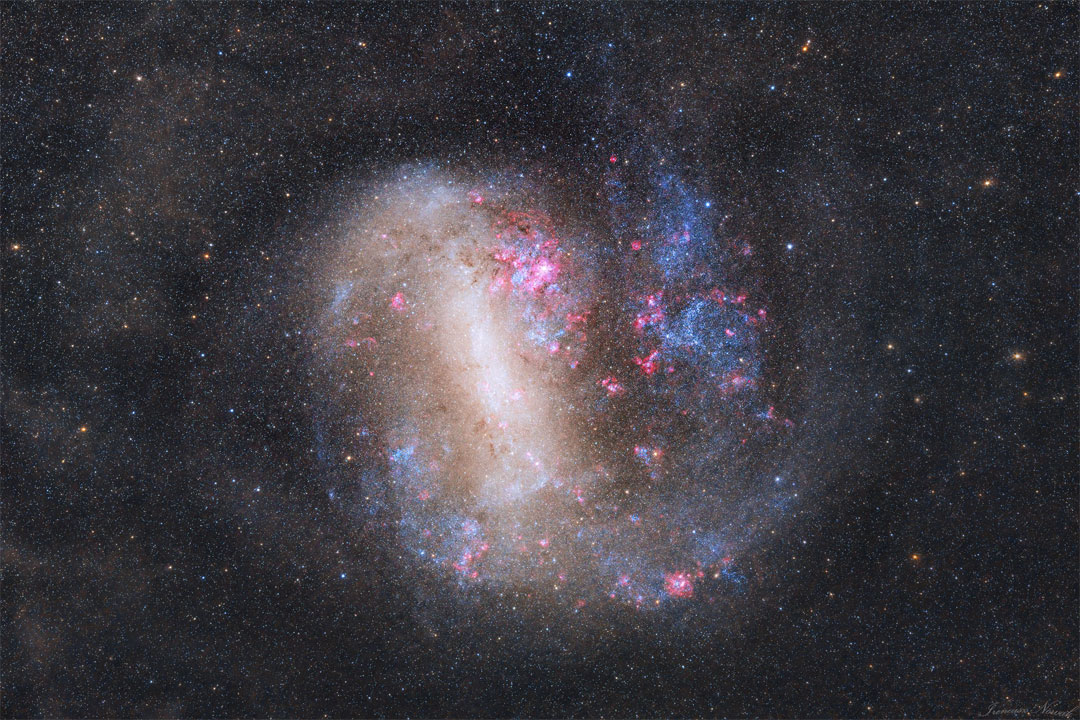 An unusual looking galaxy is shown with a light bar
running nearly vertical and blue stars and red nebulas
around the edges.
Please see the explanation for more detailed information.