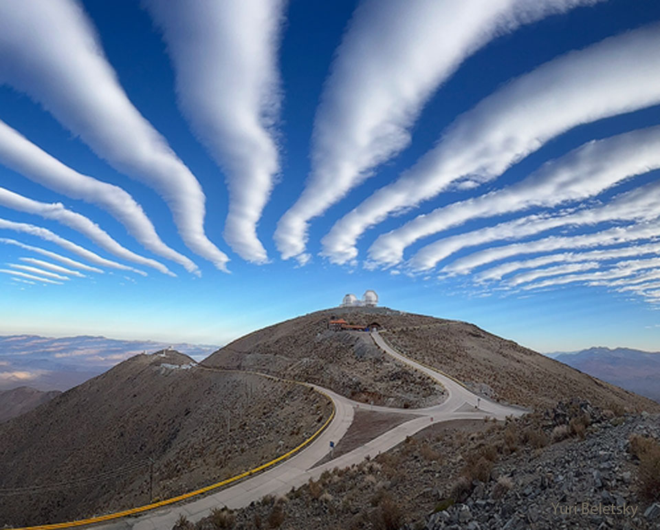 A series of white parallel clouds are seen going off
into the distance in a background blue sky. In the foreground
is a hill with two domes at the top.
Please see the explanation for more detailed information.