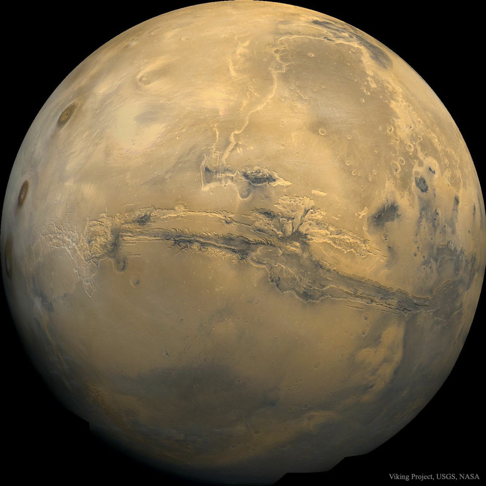 A picture of Mars is shown as a large orange globe.
Across the center of the planet a long canyon is visible.
Please see the explanation for more detailed information.