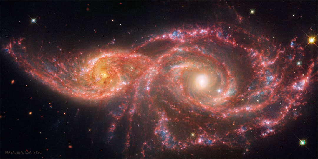 Two spiral galaxies are pictured on the left and right.
They galaxy on the left is smaller. Both show red lanes of
dust in their spiral arms.
Please see the explanation for more detailed information.