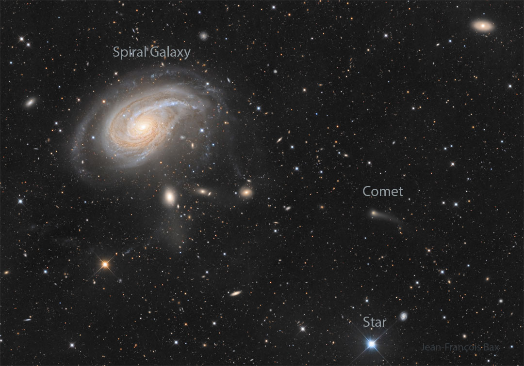 A dark field is filled with stars and galaxies. A large
spiral galaxy appears on the upper left. Toward the right,
there is a smaller fuzzy patch that is a comet with a short
tail.
Please see the explanation for more detailed information.