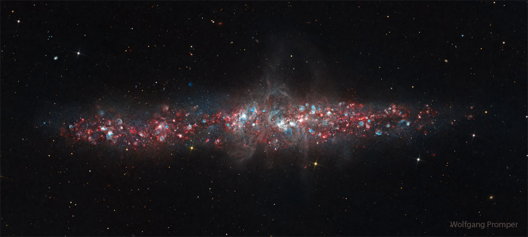 A galaxy is shown that has a lot of blue and red dots in it. The
galaxy is an long elliptical blob across the middle and not a classic spiral.
Please see the explanation for more detailed information.