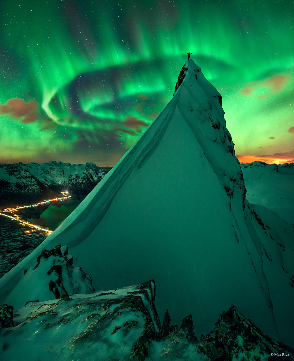 A person with the arms raised stands atop of a
rock peak covered in snow. Snow covered mountains
are all around. Green aurora swirl overhead and
reflect off the snow.
Please see the explanation for more detailed information.