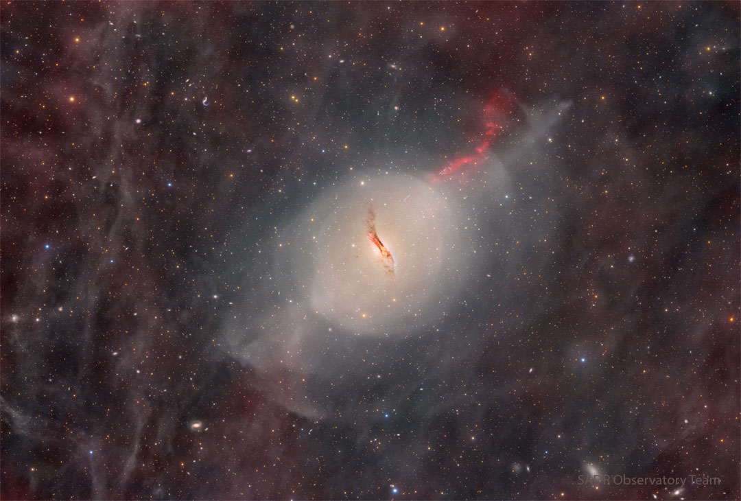 A starfield glows faintly with red glowing gas and dust.
In the center there is some sort of unusual galaxy with a red jet
emanating toward the upper right and a dark dust lane through
its center.
Please see the explanation for more detailed information.