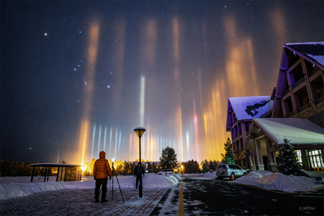 A starry sky appears above a snowy street with a
house on the right. The constellation of Orion is
visible on the left. Up from the ground many pillars
of light are seen. Two people stand watching the pillars
on the street in the foreground.
Please see the explanation for more detailed information.