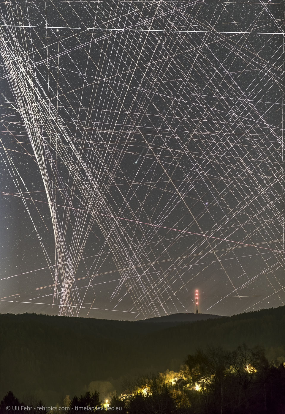 A night sky filled with lines and dashed-lines
appears above a hilly landscape featuring a distant
lit-up tower. Near the center of the image is a comet
shown by its small green coma and ion tail to the upper
right.
Please see the explanation for more detailed information.