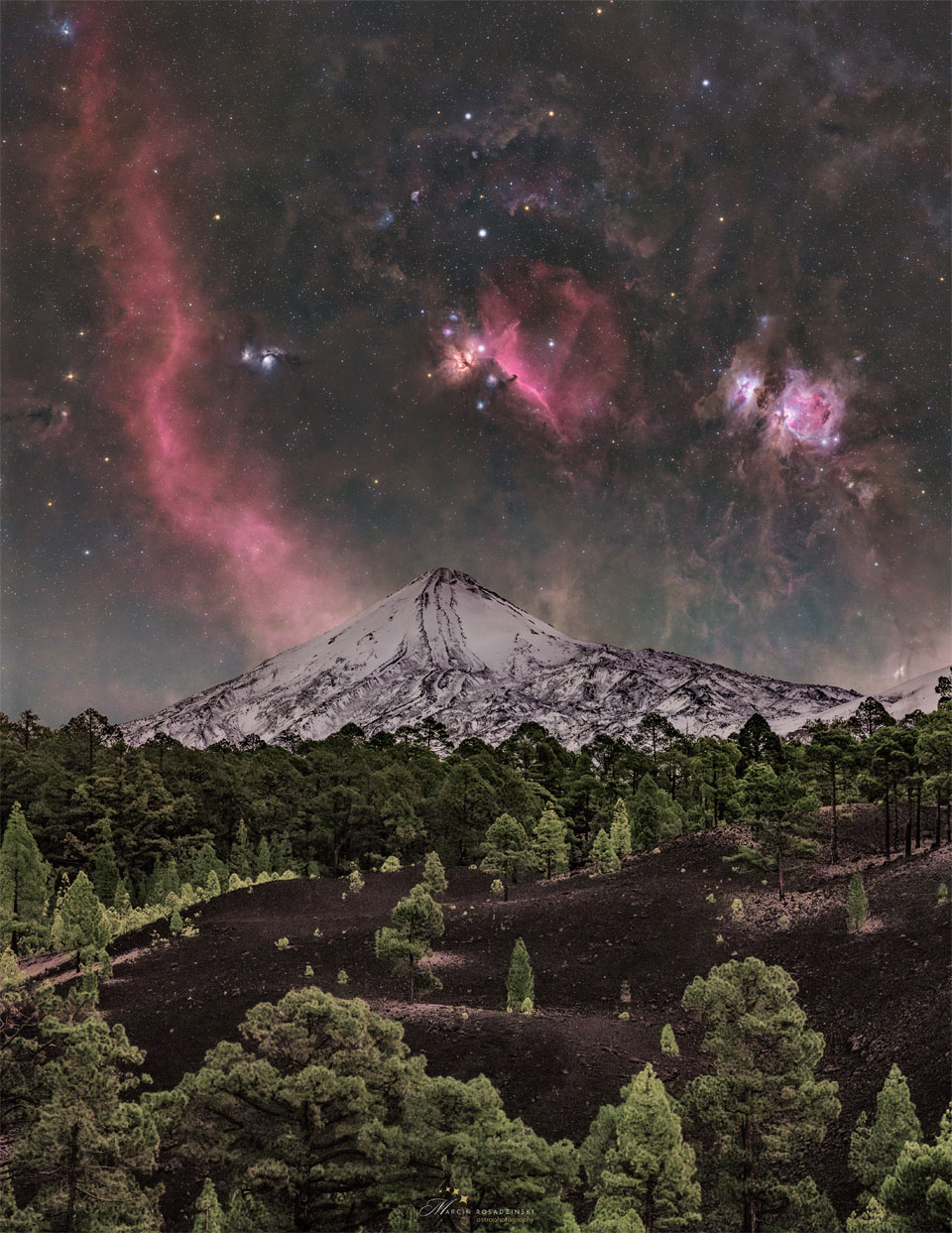 A starry sky is seen above a snowy volcano peak. The sky is
bright with many stars and red nebula. In the foreground is dark
soil and green evergreen trees.
Please see the explanation for more detailed information.
