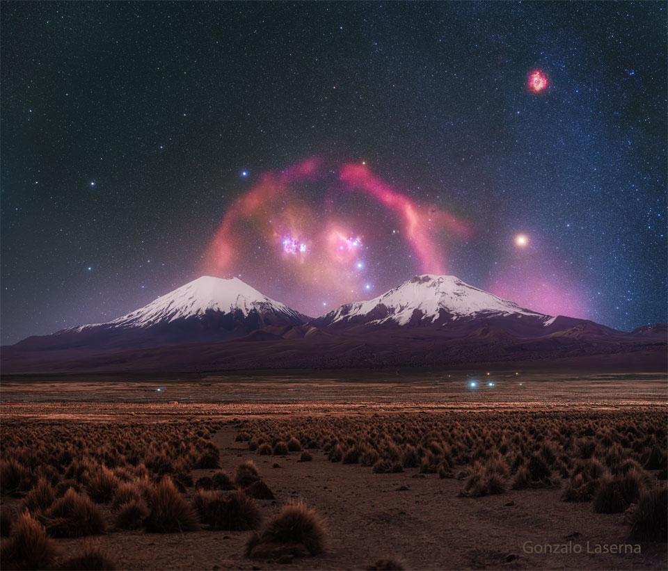 A pair of snow topped volcanoes are seen in the distance.
A background starry sky is seen behind them, with a red filamentary
nebula looping around that appears to connect the two tops
of the volcanoes.
Please see the explanation for more detailed information.