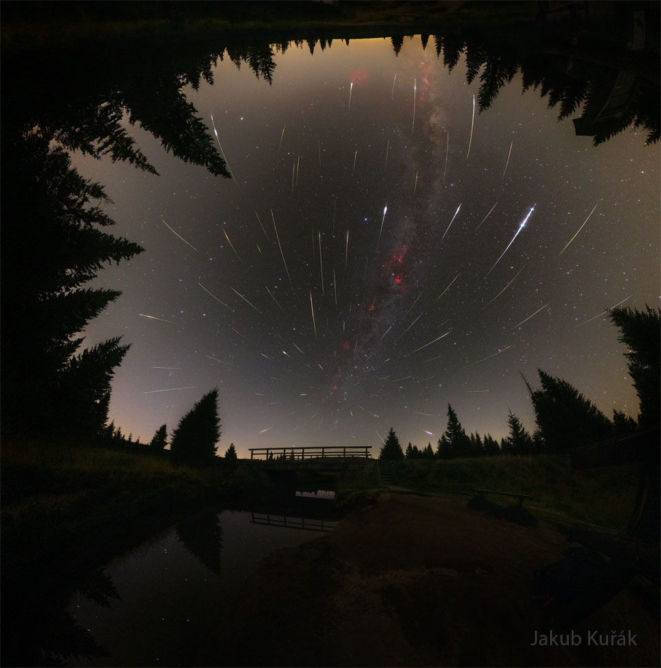 A starfield is bounded by trees on three sides and
a pond on the bottom. Amongst the stars are many streaks
of light appearing to emanate from a place on the sky
just above a foreground bridge.
Please see the explanation for more detailed information.