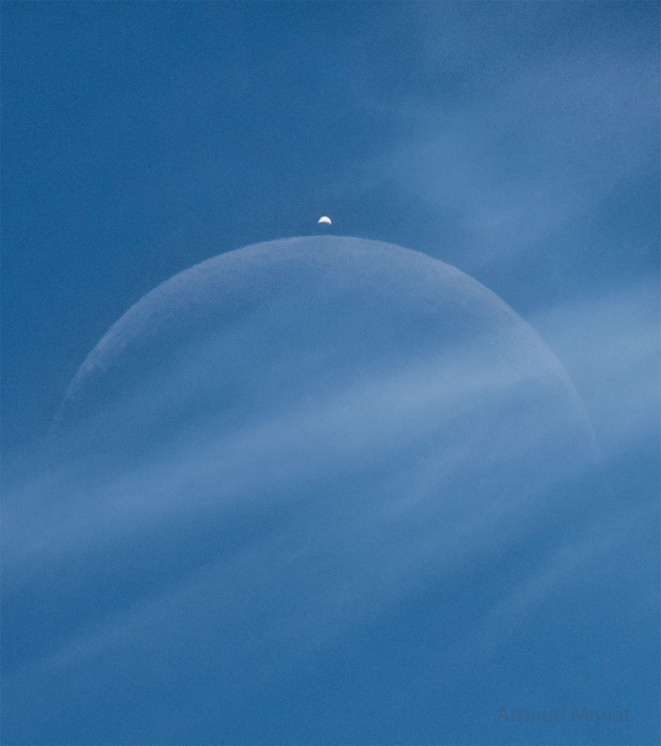A blue sky has a light cloud crossing the middle of
the image. Two crescent objects are visible through the
cloud, a large faint crescent in the center and a brighter
crescent above it.
Please see the explanation for more detailed information.