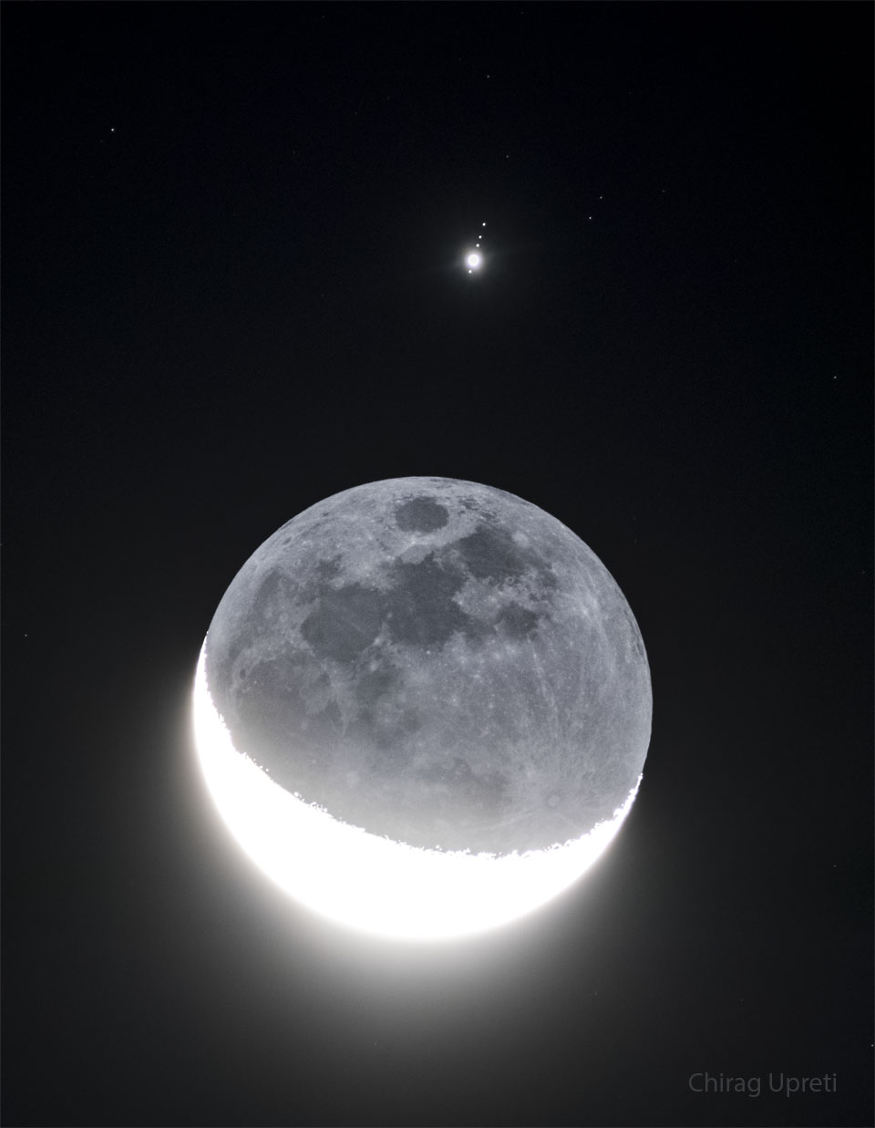 In front of a dark background an image of Earth's Moon
appears with a lower crescent brightly lit. The rest of the Moon
is also faintly visible. Above the Moon is a bright point of
light surrounded by four fainter points of light in row.
Please see the explanation for more detailed information.
